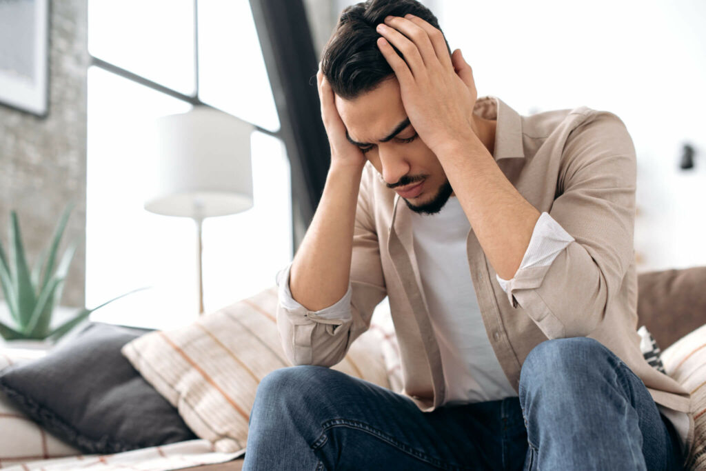 A distraught looking man sits while holding his head in his hands.